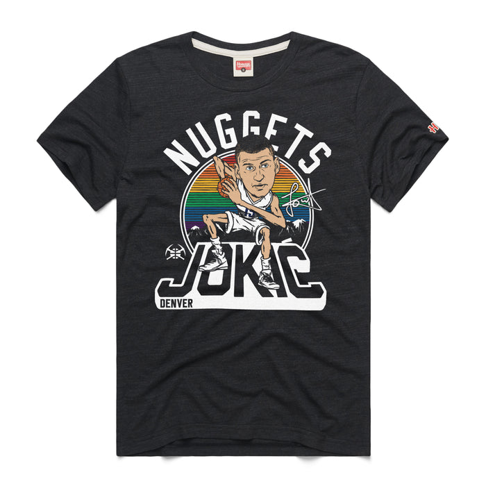 A black short-sleeve crewneck t-shirt featuring a retro graphic on the chest centered around a cartoon caricature of Nikola Joki?. Above the caricature, the word 'NUGGETS' arcs in a bold white font against a circular background with rainbow horizontal stripes and a subtle city skyline silhouette. Below, 'JOKIC' is written in a stylized, oversized 3D block font, with a small white 'DENVER' printed on the bottom left corner. A tiny red brand logo is visible on the left sleeve cuff