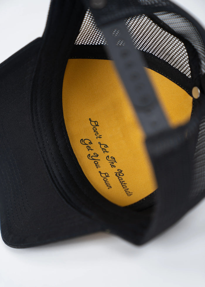 Black cap with mesh back, yellow inside reads "Don't Let The Bastards Get You Down" in elegant script.