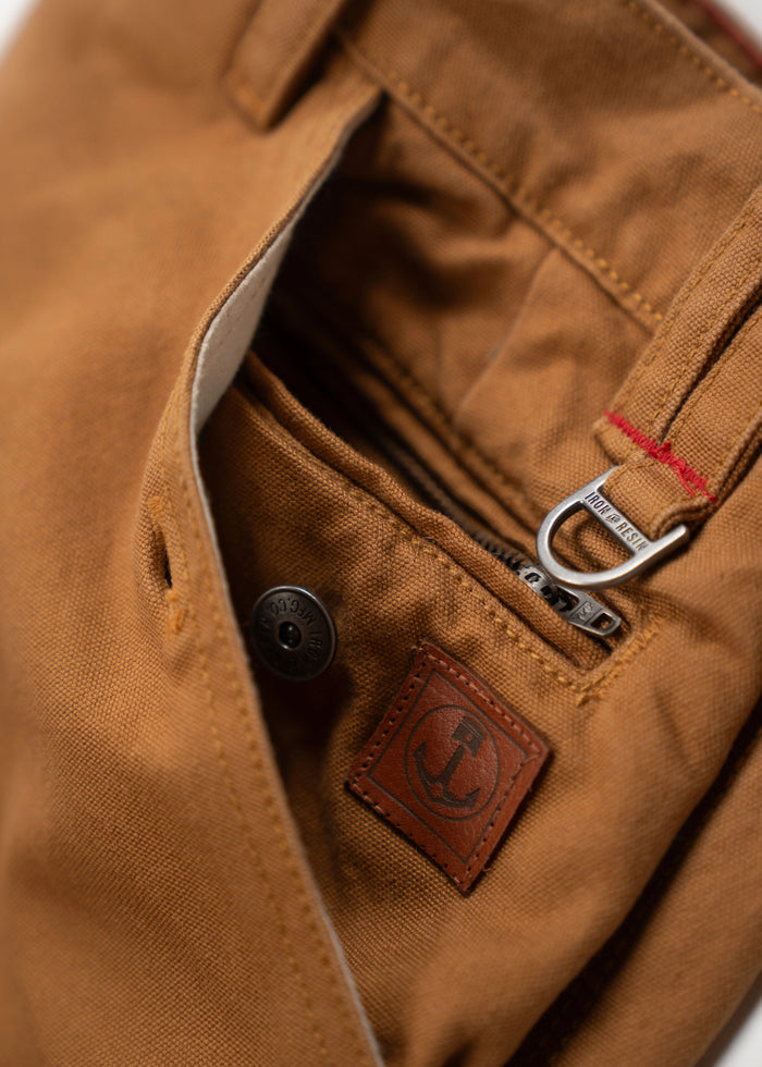 Close-up of brown pants pocket with a leather anchor patch, metal button, and zipper detail, showcasing rugged craftsmanship.