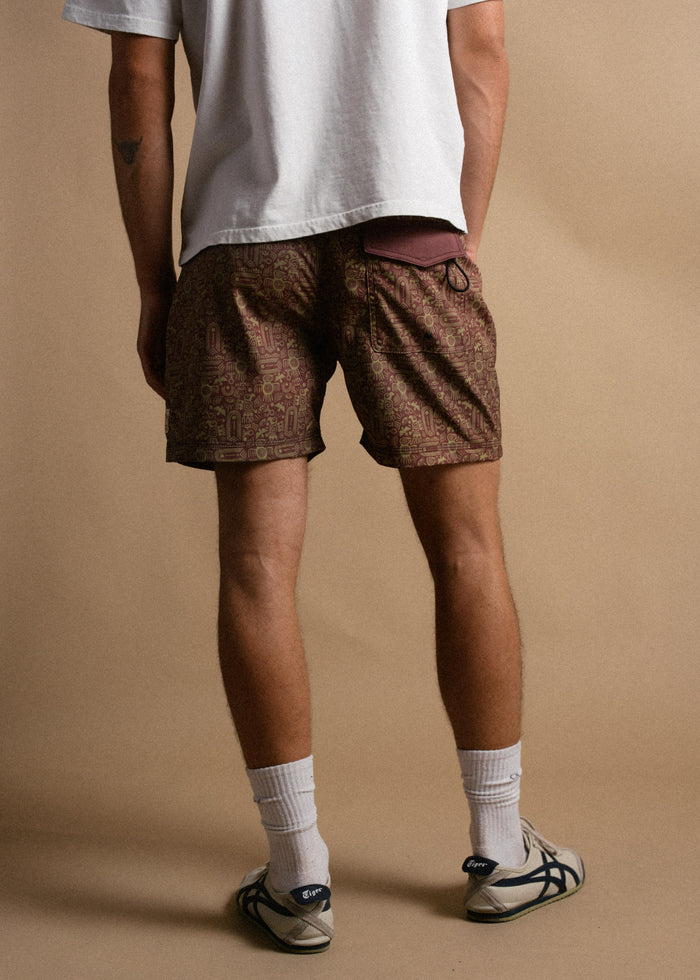 Man in a white shirt and patterned shorts stands on a beige background, wearing white socks and sneakers, facing away.