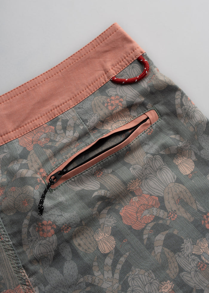 Close-up of floral-patterned shorts with a pink waistband, zipper pocket, and red loop detail on a light gray background.
