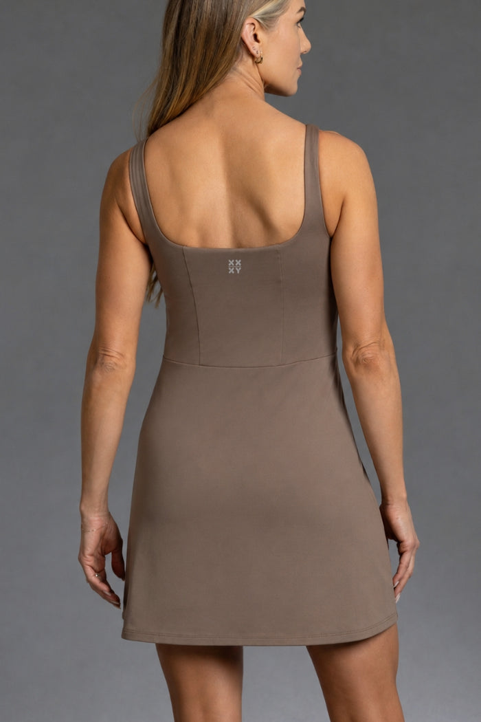 A woman shown from the back wearing a sleeveless, scoop-neck mini dress in a solid taupe-brown color, photographed against a neutral gray studio background. || mocha