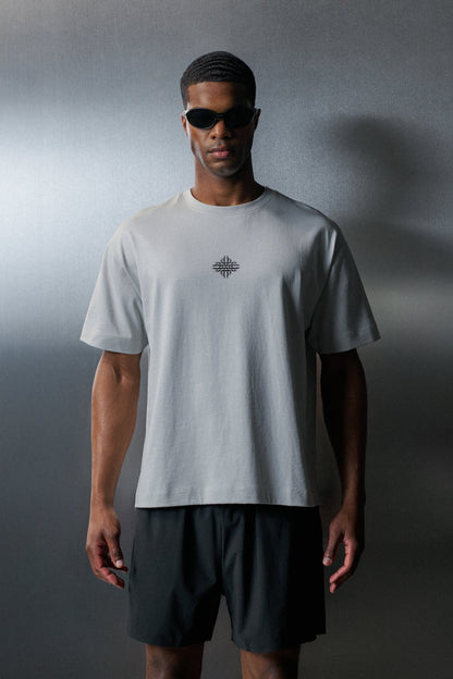 Man wearing a light grey t-shirt with a logo and dark shorts against a gradient grey background