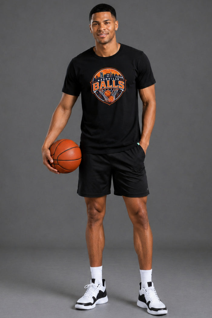 A man modeling a black short-sleeve graphic t-shirt with an orange and blue Phoenix Suns logo, paired with black athletic shorts, white socks, and black-and-white sneakers, holding a basketball against a solid gray studio background. || Black