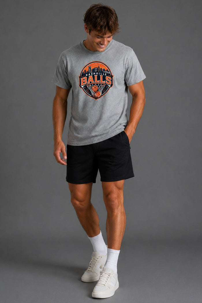 A man modeling a heather gray t-shirt with a 'Falcon Bats' graphic on the chest, paired with black athletic shorts, white socks, and white sneakers against a solid gray studio background. || Heather Gray