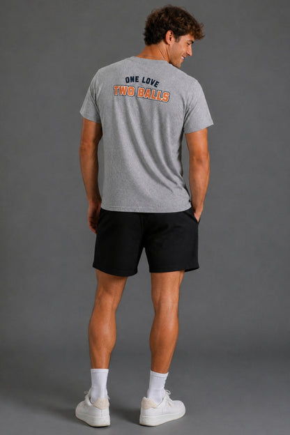 A man wearing a heather gray t-shirt with the text 'ONE LOVE TWO BALLS' printed on the back, paired with black athletic shorts, white socks, and white sneakers, photographed from behind against a solid gray studio background. || Heather Gray