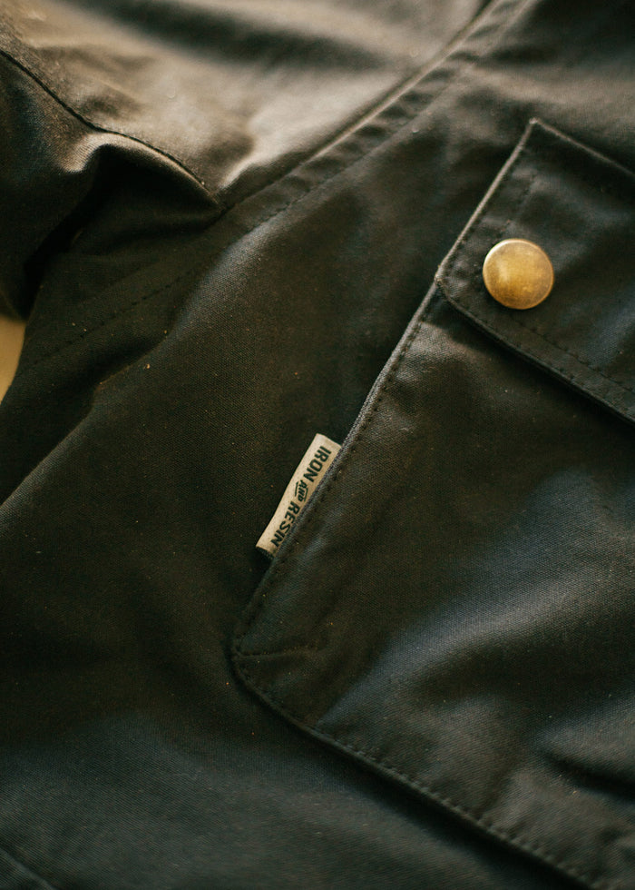 Close-up of a dark fabric jacket with a brass button and visible "Iron & Resin" label near the pocket, highlighting details.