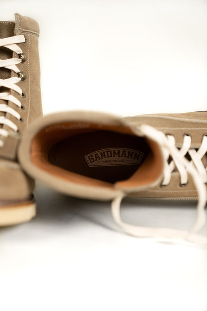 Tan suede boots with white laces, one tilted showing "Sandmann Handcrafted Goods" logo inside, on a light background.