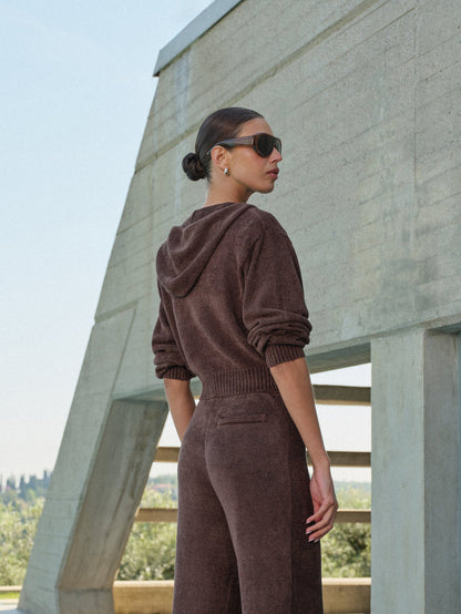 Woman in a brown matching tracksuit standing against a concrete structure with a clear sky.